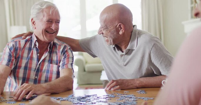 Two diverse senior male friends embracing, doing jigsaw puzzle in living room, slow motion