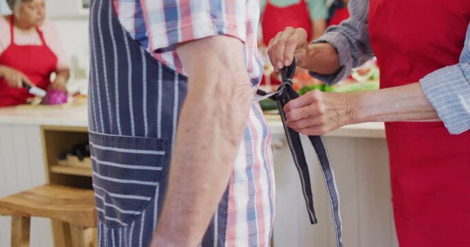 Happy caucasian senior couple putting on aprons, in kitchen with diverse friends, slow motion