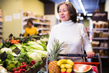 Obraz premium Portrait of thoughtful elderly woman walking among shelves with products in grocery shop, carrying shopping trolley..
