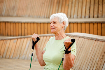 Old woman with short hair doing exercises outdoor , sitting on wooden bench using resistance rubber bands.