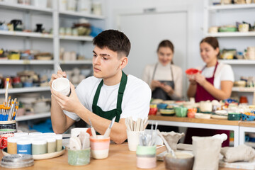 Young man painting clay ceramic cup in workshop