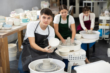 Joyous young man in pinafore holding handmade clay dish sitting at potter wheel in art studio.