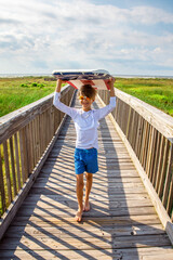 Young Preteen Boy Wearing Sunglasses and Walking with Boogie Board Over His Head On A Boardwalk