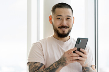  Smiling Asian man with stylish tattoos holding smartphone using mobile app shopping online at home. Modern bearded hipster ordering food. Mobile banking, technology concept 