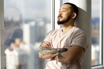 Asian man with arms crossed, closed eyes close-up standing near window, thinking about something. A...