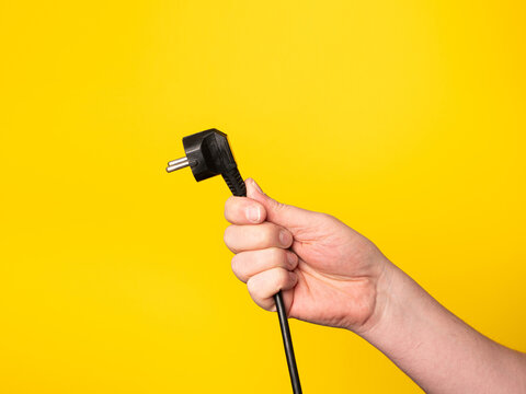 A hand holds a plug to which a black cable is attached.  No face, yellow background.
