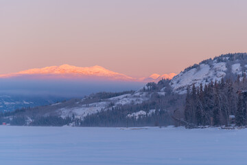 Landscape winter views in northern Canada during sunrise with pink tones on surrounding mountains and sky. Frozen lake in foreground. 