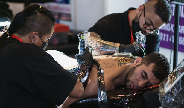two tattoo artists working at the same time on the back of a man lying down with an expression of pain, use of biosecurity, art fusion