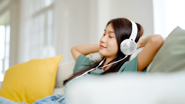 Young asian woman listening to music on couch in living room at home. Happy asia female using mobile smartphone, wearing headset and sitting on sofa