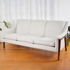 Vintage white sofa. Stylish mid-century modern couch with sculptural wooden legs. Interior scene with white curtains and wooden floor.