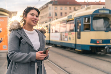 Smiling young asian woman checking schedule table of city traffic online on application using smartphone,asia woman satisfied use app for paying electric transport via cellphone waiting for tram