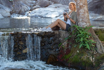 Female hiker looking at waterfall, Happy female backpacker enjoying amazing tropical waterfall, traveller or Inspiring travel blogger make photos for social media on beautiful tropical waterfall.