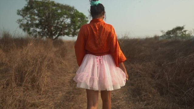 A slow motion shot of the back of an outgoing carefree Asian female wearing a pink tutu dress, stretching her arms out while walking along a pathway in a dry grass field, India 