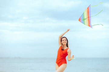 Happy Woman Holding a Rainbow Kite at the Beach. Beautiful lady feeling carefree and relaxed wearing a swimsuit
