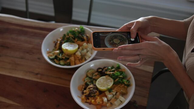 Woman takes photos of delicious vegan vegetarian meal with phone on dinner table - close up on hands and smartphone
