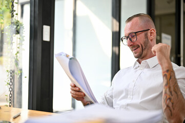 Fototapeta premium Happy Caucasian businessman looking at business paperwork, showing yes gesture. good news