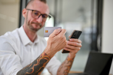 Obraz na płótnie Canvas Thoughtful Caucasian man with glasses and tattoo on his arms holding a credit card and his phone