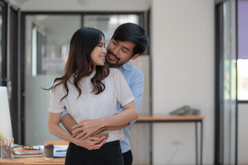Attractive smiling Asian man hugging woman indoors. Beautiful successful couple embracing the workplace. Husband and wife hug togetherness concept.