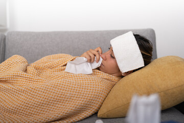 Young woman with fever blowing her nose, while holding a wet towel on her forehead