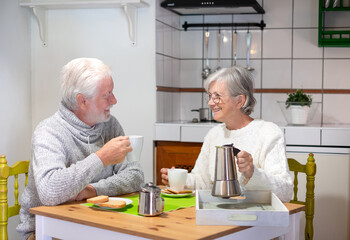 Smiling senior couple having breakfast together at the table at home. Elderly wife holding a coffee-maker, old people enjoying retirement
