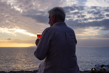 Rear view of senior man sitting in front of the sea at sunset drinking a glass of wine, horizon over the water. Lonely old man drowns his problems in alcohol