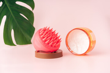 Head massager and head cream against pink background and monstera leaf.
