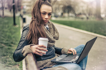 Long hair hipster brunette woman sitting on a bench in the park,  working on a laptop and drinking a cup of coffee.