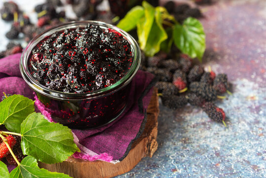 homemade mulberry jam in a glass jar with fresh mulberry with green leaves on rustic table background.