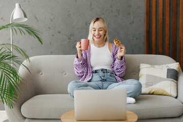 Young woman wear casual clothes watch movie film on laptop pc computer drink coffee eat cookie sits on grey sofa couch stay at home hotel flat rest relax spend free spare time in living room indoor.