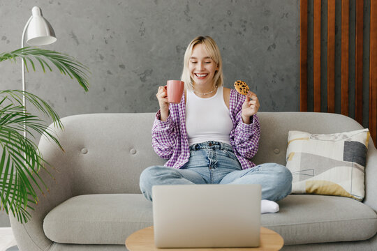 Young woman wear casual clothes watch movie film on laptop pc computer drink coffee eat cookie sits on grey sofa couch stay at home hotel flat rest relax spend free spare time in living room indoor.