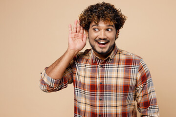 Curious nosy smiling Indian man wear brown shirt casual clothes try to hear you overhear listening intently isolated on plain pastel light beige background studio portrait. People lifestyle concept.