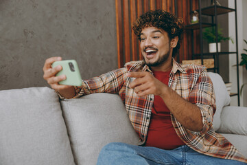 Young Indian man wear casual clothes use play racing app on mobile cell phone gadget for pc video games sits on grey sofa couch stay at home hotel flat rest relax spend free spare time in living room