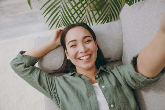 Close up young woman of Asian ethnicity wears casual clothes doing selfie shot pov on mobile cell phone sits on grey sofa couch stay at home hotel flat rest relax spend free spare time in living room.