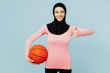 Young arabian muslim fitness trainer sporty woman wear pink abaya hijab spend time home gym hold basketball ball show thumb up isolated on plain blue background studio Workout sport fit abs concept