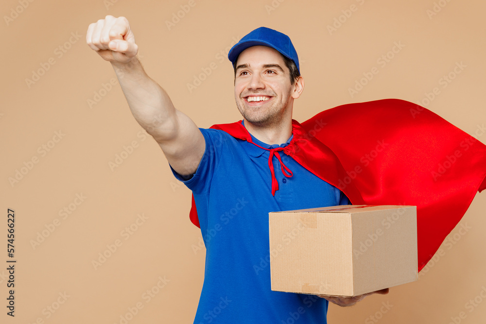 Wall mural Delivery guy employee man wearing blue cap t-shirt uniform workwear super hero red coat work as dealer courier hold cardboard box do flying gesture isolated on plain beige background. Service concept.