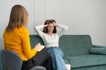 Young woman at a psychologist's appointment is sitting on the couch and holding her head,...