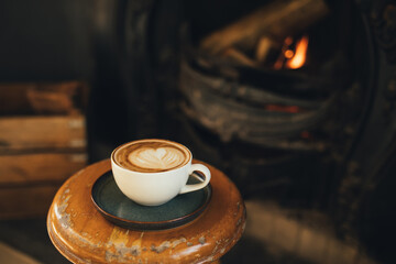 Cup of hot cappuccino on wooden stool near burning fireplace.