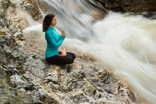 Woman practicing yoga in lotus position on rock by rushing river, Gorham, New Hampshire, USA