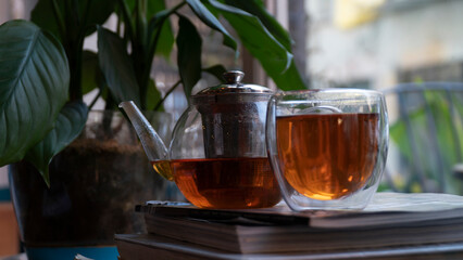 Red rooibos tea in a pot and in a glass mug on the magazine pile. There is a window and plants too.