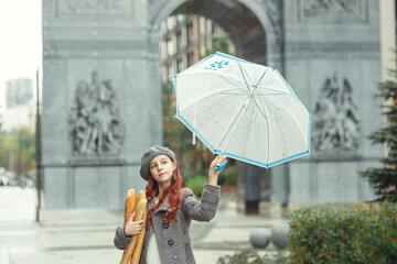 Beautiful stylish slim girl in elegant coat and beret holding wet umbrella and baguettes at city...