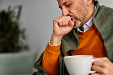 Close-up of a sick senior man, coughing and holding a cup of tea at home.
