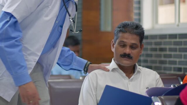 A helpful and supportive Indian Asian male doctor wearing an apron and stethoscope showing a file or reports talking and consoling a sad and worried patient's relative sitting a hospital waiting area