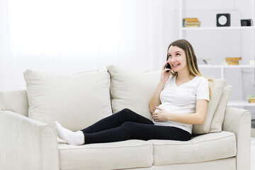 Smiling pregnant woman talking over the phone on white sofa.