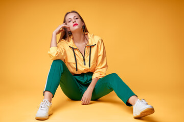 Colorful portrait of thoughtful young girl in sports casual wear sitting on yellow background.