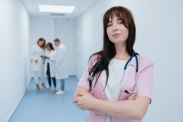 Successful nurse looks into the camera against the background of colleagues who are standing in the corridor of the clinic. Meeting on the go.