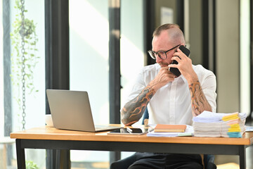 Handsome adult man employee in white shirt and glasses having phone conversation while sitting at desk in modern office