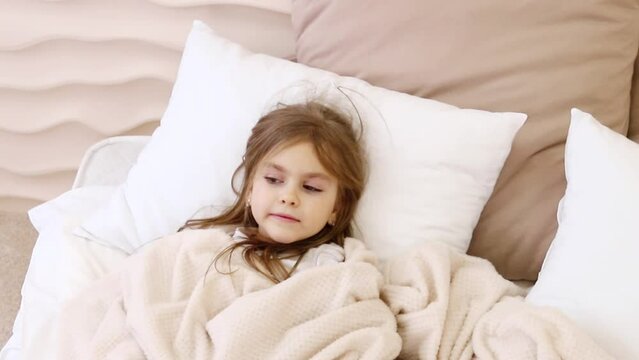 Child wake up. Little girl lies in bed under blanket early morning with shaggy hair on face hard wake up.