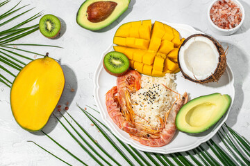 tropical bowl with avocado, prawns, rice, mango, kiwi and coconut, tropical food on the sea beach on light background with tropical leaves. Healthy food, clean eating, Buddha bowl, top view