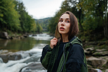 Portrait of a beautiful woman in a jacket while traveling on the background of a river and mountain landscape, hiking in the mountains, looking away with a serious face