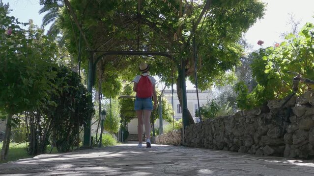Traveler tourist woman with hat and red backpack explore the gardens of Achillion palace on Corfu island, Greece, slow motion